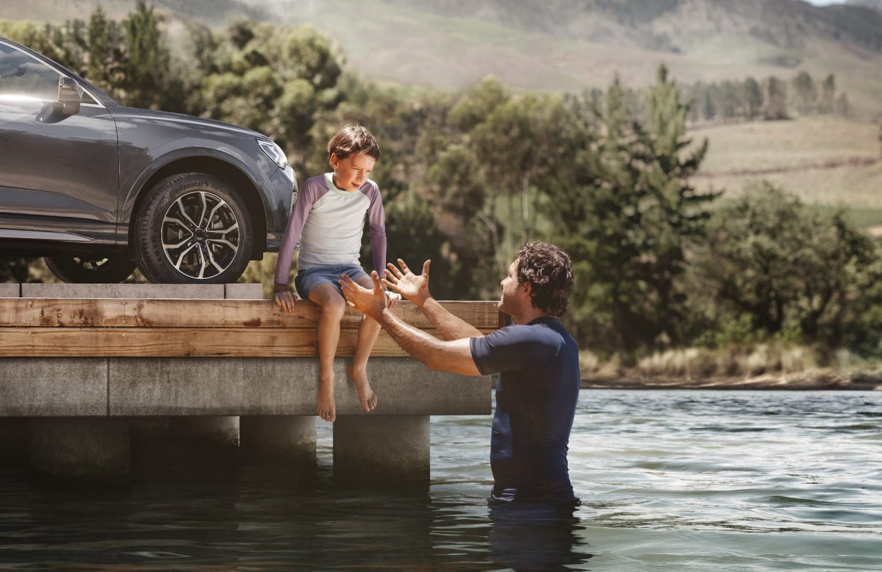 Ein Kind sitzt auf einem Steg, während der Vater im Wassers steht und die Hände zu dem Kind als Hilfestellung ausstreckt. Im Hintergrund steht ein Auto auf dem Steg mit Continental Reifen..