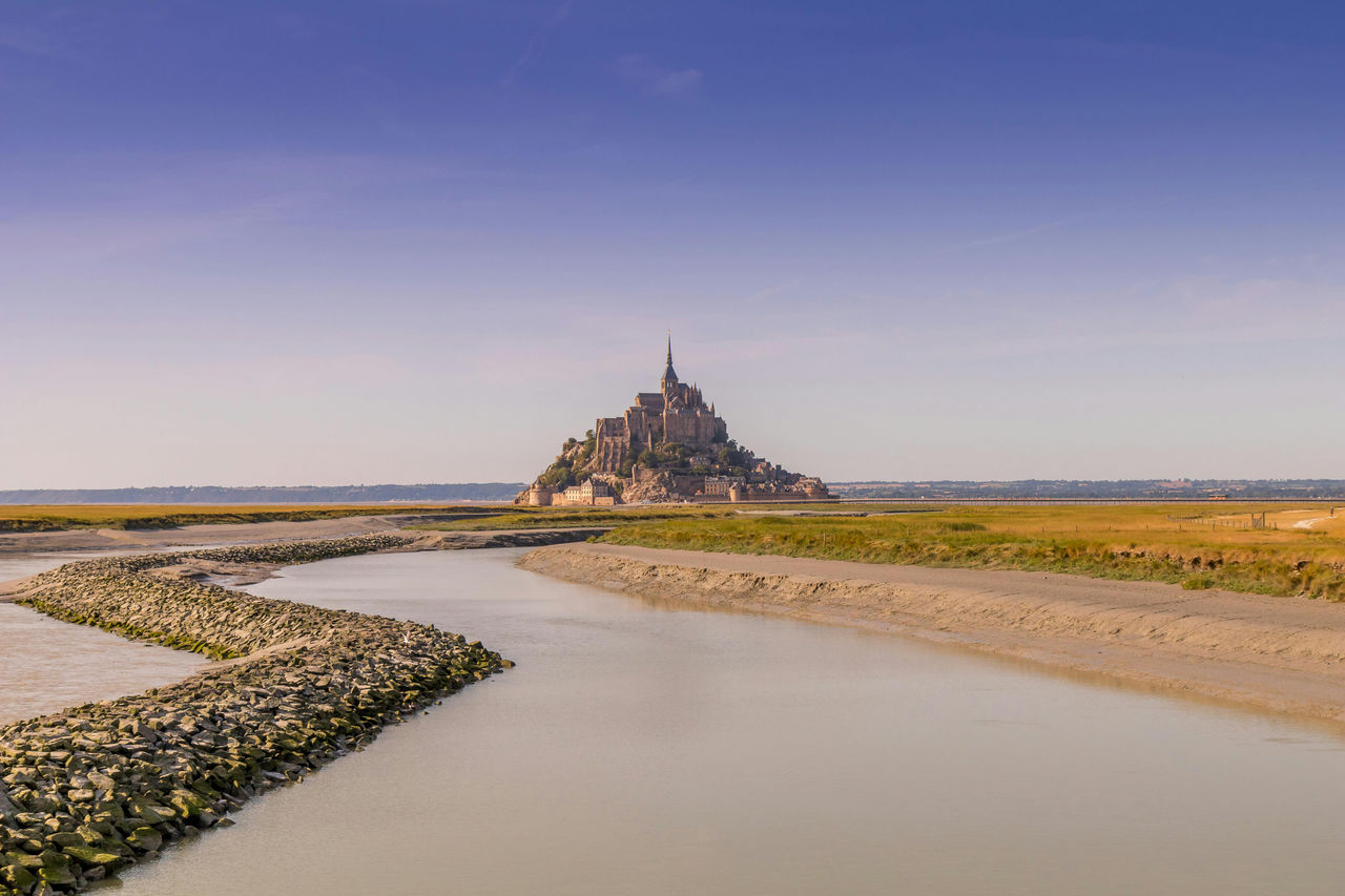 Der Mont-Saint-Michel erhebt sich aus den Wattflächen am Rande der Normandie und der Bretagne, umrahmt von einem ruhigen Fluss und weitem Himmel.