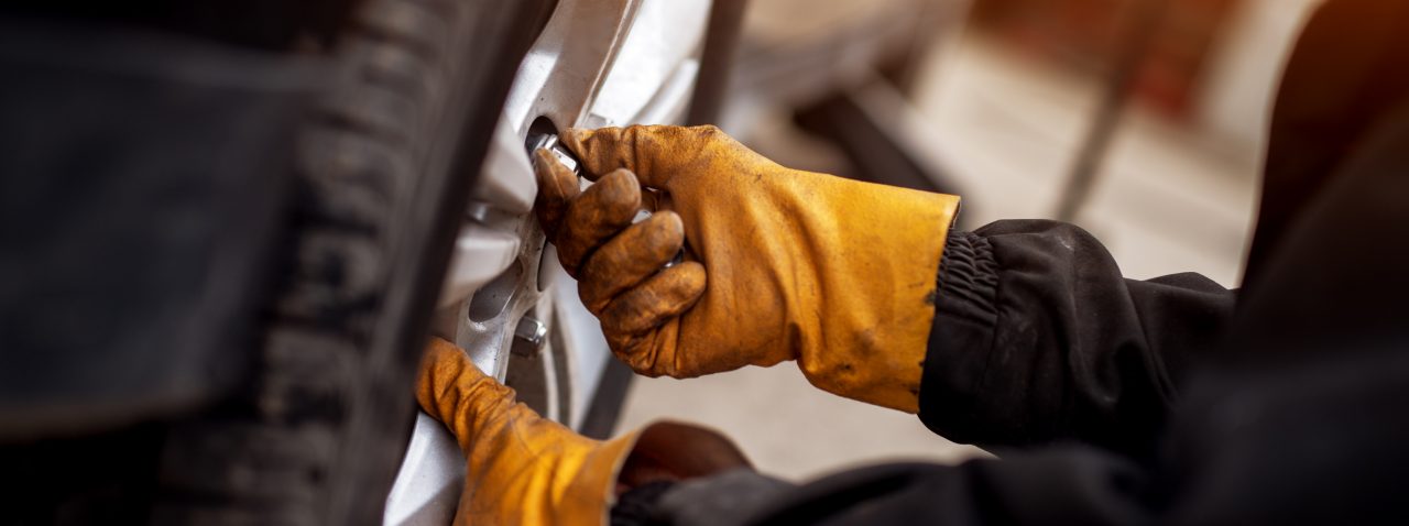 An experienced mechanic with orange gloves is putting screws on a placed wheel on a car.