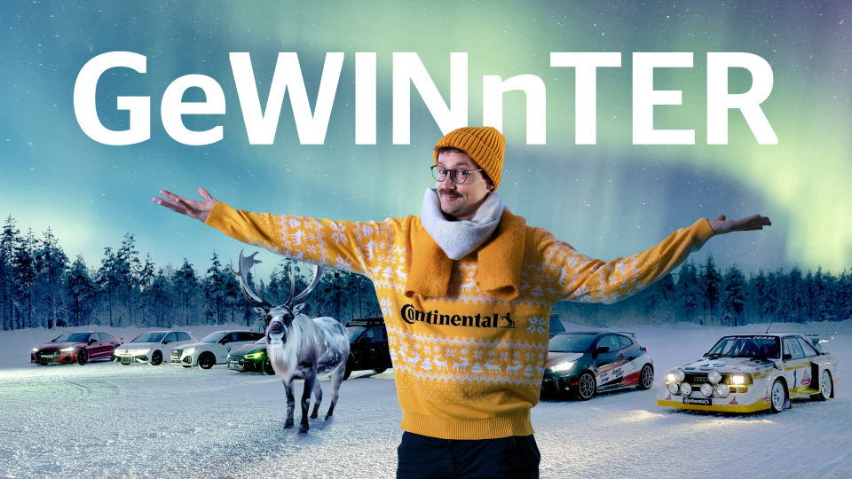 Mann in gelber Continental-Kleidung vor verschneiter Landschaft mit Polarlichtern, Autos und Rentier im Hintergrund.