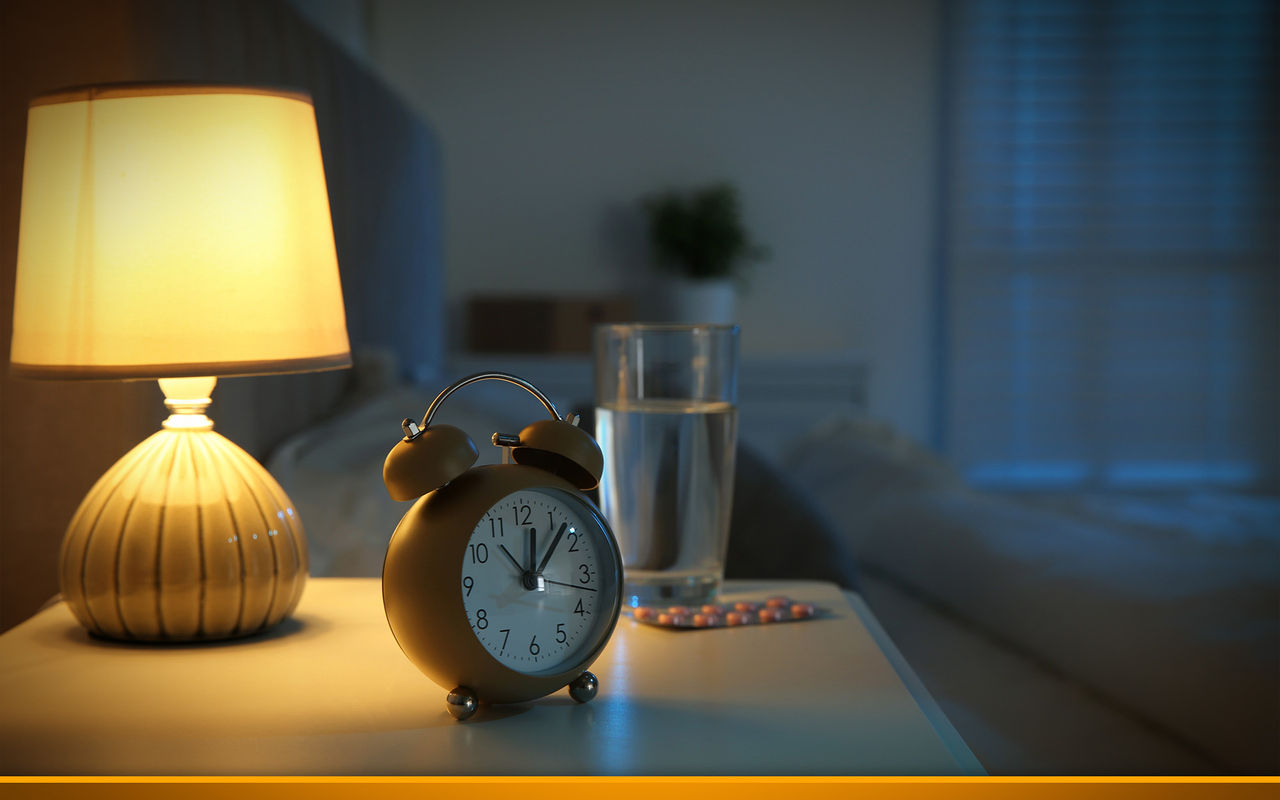 Insomnia treatment. Alarm clock, glass of water and pills on bedside table in bedroom at night